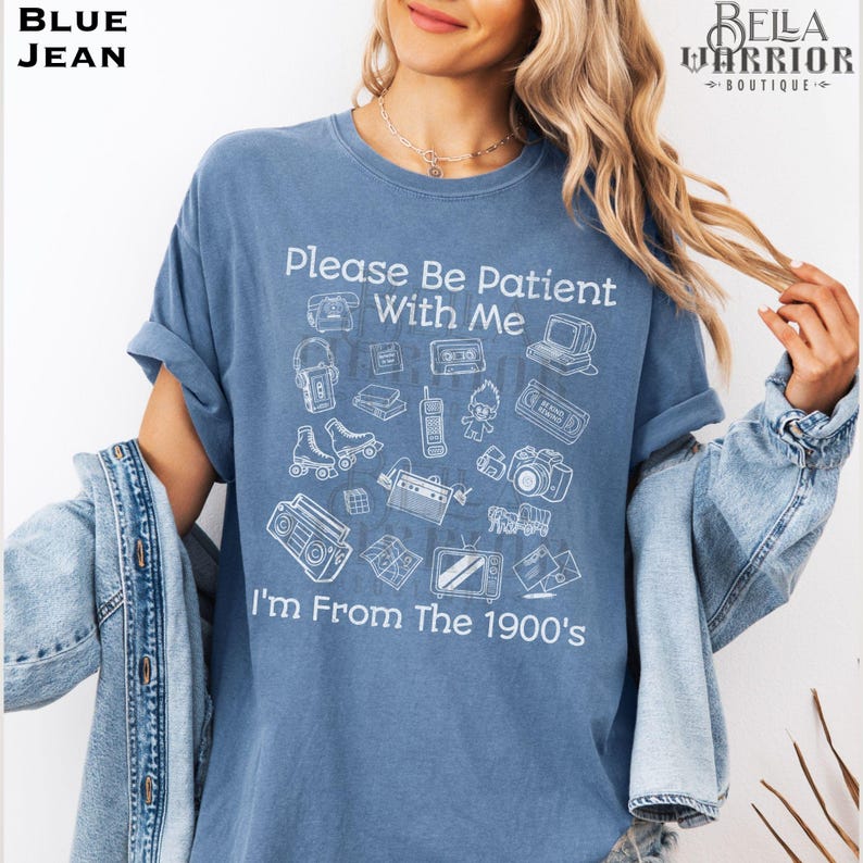 Puede incluir: Una camiseta color azul jean con gr&aacute;ficos de l&iacute;neas blancas de art&iacute;culos vintage que incluyen un tel&eacute;fono de disco, un casete, un radiocasete y un televisor. El texto dice, Please Be Patient With Me I'm From The 1900's, en fuente blanca.
