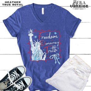 May include: Heather true royal V-neck t-shirt with a graphic of the Statue of Liberty and the words "Freedom, Democracy, Truth". The shirt is paired with white jeans and black sneakers.