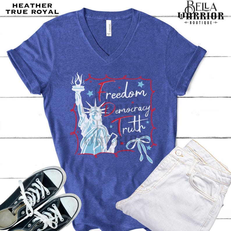 May include: Heather true royal V-neck t-shirt with a graphic of the Statue of Liberty and the words "Freedom, Democracy, Truth". The shirt is paired with white jeans and black sneakers.