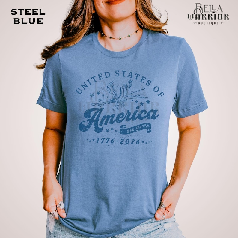 Puede incluir: Camiseta azul acero con un gr&aacute;fico desgastado que dice "United States of America" con un &aacute;guila y estrellas. Tambi&eacute;n incluye el texto "250 Years" y las fechas "1776-2026". La camiseta es de cuello redondo.