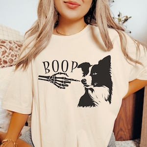 May include: A white t-shirt featuring a black and white graphic of a border collie with a skeleton hand reaching out to boop the dog's nose. The text "BOOP" is above the graphic.