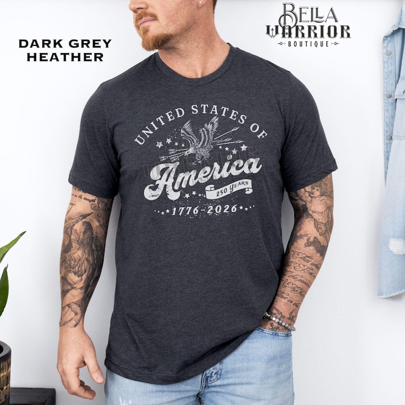 Puede incluir: Camiseta de color gris oscuro jaspeado con un gr&aacute;fico blanco desgastado que dice "United States of America" con un &aacute;guila y los a&ntilde;os "1776-2026". La camiseta es de manga corta y tiene cuello redondo.