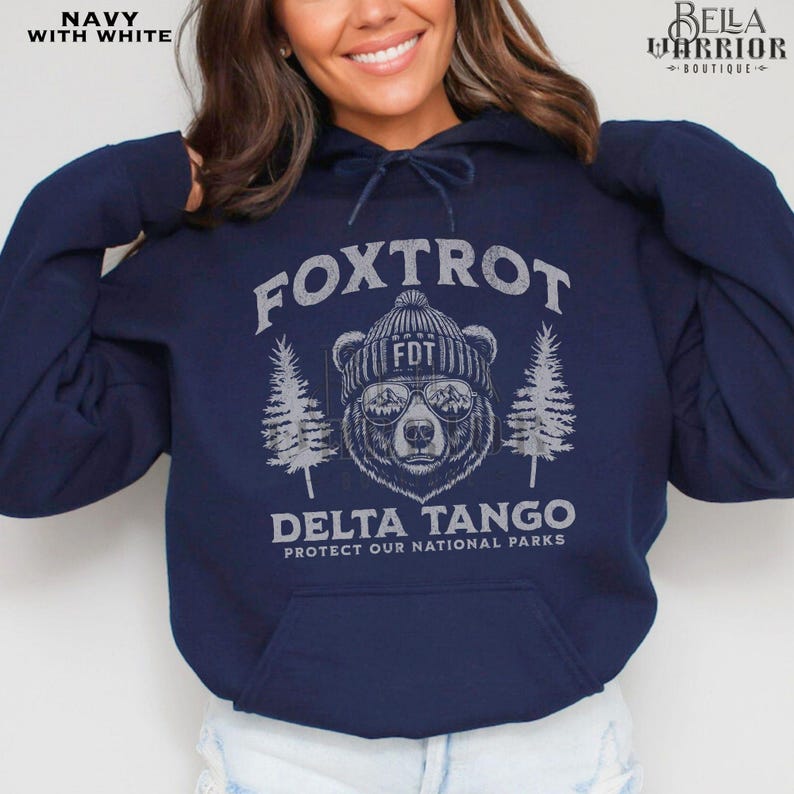 Puede incluir: Sudadera con capucha azul marino con un gr&aacute;fico blanco de un oso con un sombrero y gafas de sol. El texto "Fox Trot Delta Tango Protect Our National Parks" est&aacute; impreso en la sudadera.