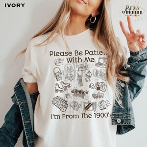 Puede incluir: Una camiseta de color marfil con ilustraciones de l&iacute;neas marrones de art&iacute;culos de los a&ntilde;os 90, incluyendo un reproductor de casetes, patines, un radiocasete y un mu&ntilde;eco troll. El texto dice, Please Be Patient With Me I'm From The 1900's.