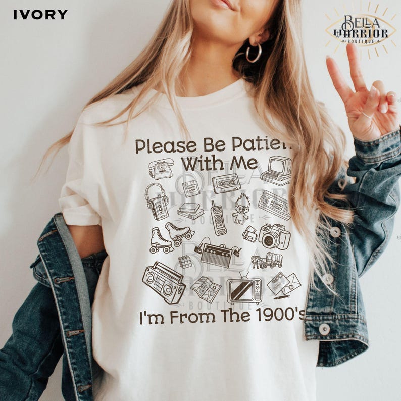 Puede incluir: Una camiseta de color marfil con ilustraciones de l&iacute;neas marrones de art&iacute;culos de los a&ntilde;os 90, incluyendo un reproductor de casetes, patines, un radiocasete y un mu&ntilde;eco troll. El texto dice, Please Be Patient With Me I'm From The 1900's.