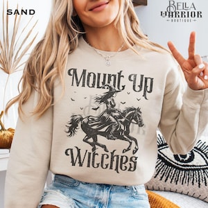 Könnte beinhalten: Sandfarbenes Sweatshirt mit dem Text "Mount Up Witches" und einer Grafik einer Hexe auf einem Pferd. Das Design ist schwarz und zeigt Fledermäuse. Das Logo von Bella Warrior Boutique befindet sich oben rechts.