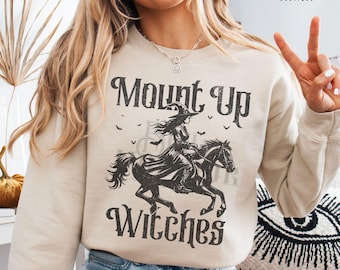 May include: Sand-colored sweatshirt with the text "Mount Up Witches" and a graphic of a witch riding a horse. The design is in black and features bats. The Bella Warrior Boutique logo is in the upper right corner.