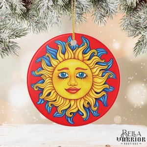 Może przedstawiać: Okrągła, czerwona ceramiczna ozdoba przedstawiająca uśmiechnięte słońce z żółtymi promieniami i niebieskimi akcentami. Słońce ma niebieskie oczy i czerwone usta. Do zawieszenia przymocowany jest złoty sznurek. Ozdoba jest przedmiotem dekoracyjnym.