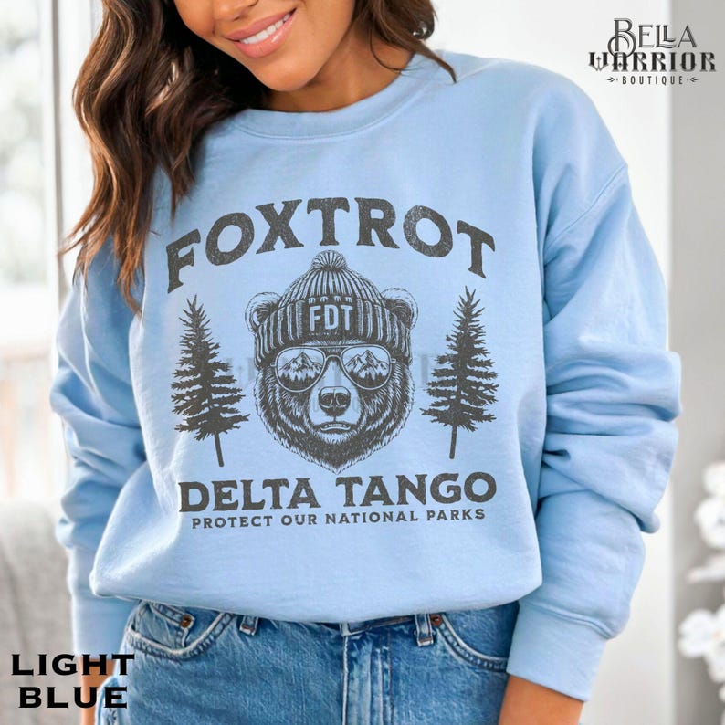 Puede incluir: Sudadera azul claro con un oso negro que lleva gafas de sol y un gorro que dice "FDT". El texto "Fox Trot Delta Tango Protect Our National Parks" est&aacute; impreso en la sudadera.