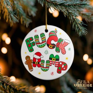 Puede incluir: Un adorno navideño de cerámica blanca con las palabras "FUCK TRUMP" en letras festivas y coloridas. Las letras están decoradas con motivos navideños, incluyendo hombres de jengibre, gorros de Papá Noel y árboles de Navidad. El adorno cuelga de una cuerda dorada.