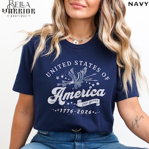 Puede incluir: Camiseta azul marino con un dise&ntilde;o gr&aacute;fico blanco. El dise&ntilde;o presenta un &aacute;guila, estrellas y el texto "UNITED STATES OF AMERICA 250 YEARS 1776-2026". El logotipo de Bella Warrior Boutique est&aacute; en la esquina superior izquierda.