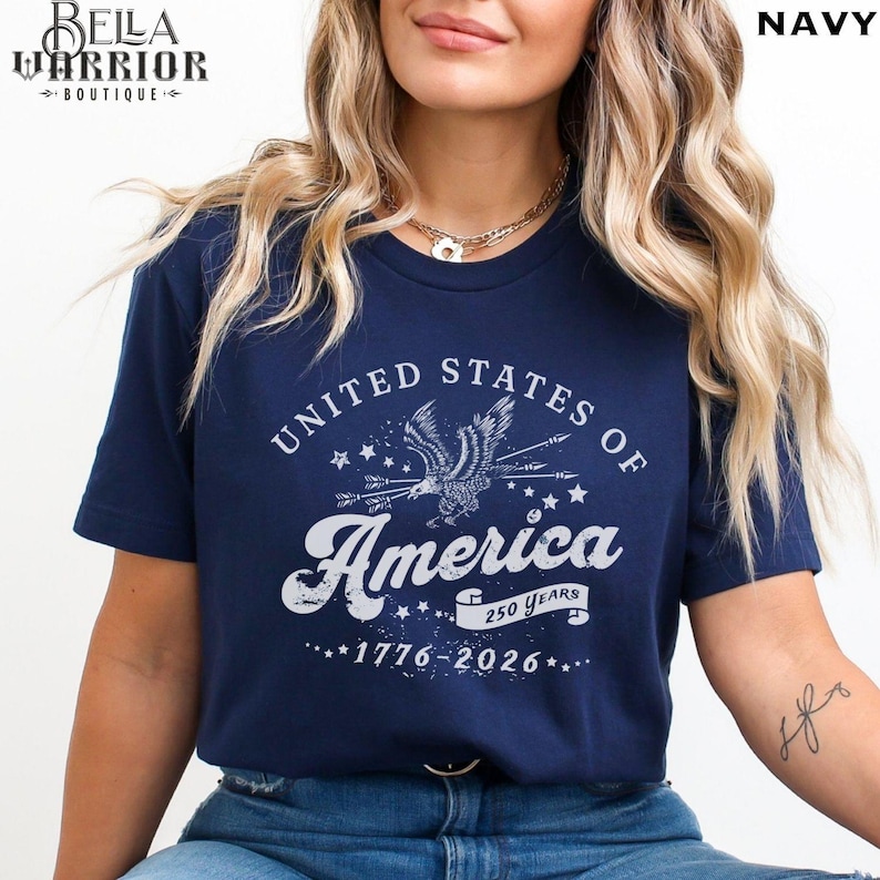 Puede incluir: Camiseta azul marino con un dise&ntilde;o gr&aacute;fico blanco. El dise&ntilde;o presenta un &aacute;guila, estrellas y el texto "UNITED STATES OF AMERICA 250 YEARS 1776-2026". El logotipo de Bella Warrior Boutique est&aacute; en la esquina superior izquierda.