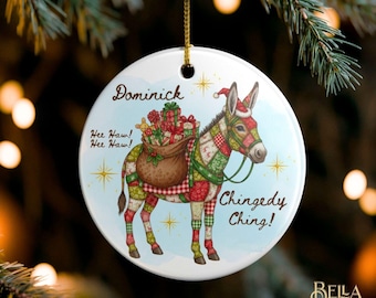 May include: A round white ceramic ornament featuring a donkey with a patchwork pattern, wearing a Santa hat and carrying a brown sack of gifts. The text reads Dominick, Hee Haw! Hee Haw!, and Chingedy Ching! with gold star accents.