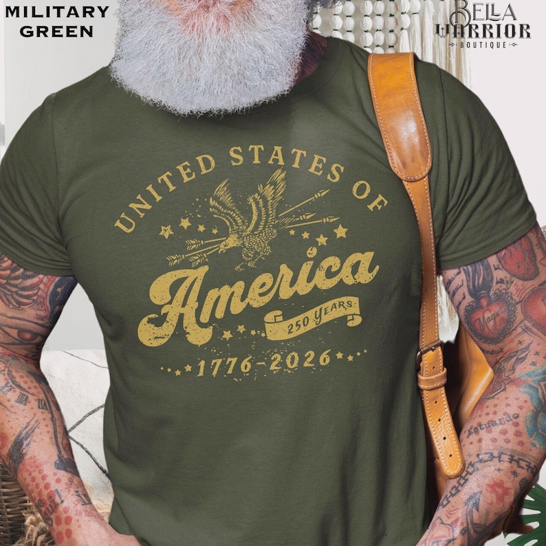 Puede incluir: Camiseta verde militar con un gr&aacute;fico dorado. El gr&aacute;fico dice "United States of America" con un emblema de &aacute;guila y "250 Years 1776-2026". La camiseta es de cuello redondo y tiene mangas cortas.