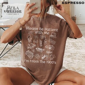 Puede incluir: Una camiseta marr&oacute;n de manga corta con texto blanco que dice Please Be Patient With Me I'm From The 1900's. El gr&aacute;fico incluye ilustraciones de l&iacute;nea de art&iacute;culos vintage como un reproductor de casetes, patines, un radiocasete y una c&aacute;mara.