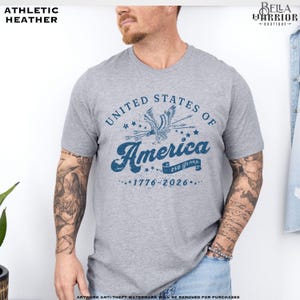 Puede incluir: Camiseta gris jaspeado con un gr&aacute;fico azul que dice "United States of America" con un &aacute;guila y los a&ntilde;os "1776-2026". La camiseta tiene cuello redondo y mangas cortas.