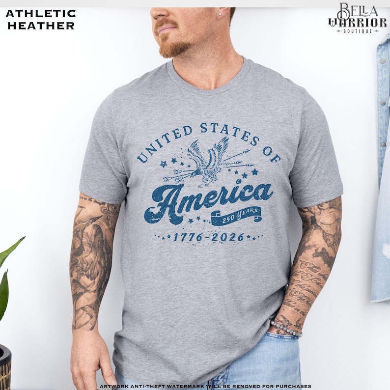 Puede incluir: Camiseta gris jaspeado con un gr&aacute;fico azul que dice "United States of America" con un &aacute;guila y los a&ntilde;os "1776-2026". La camiseta tiene cuello redondo y mangas cortas.