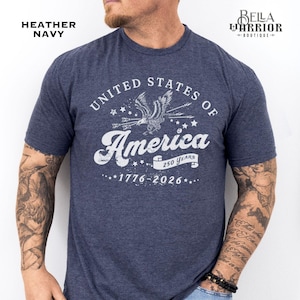 Puede incluir: Camiseta azul marino jaspeada con un dise&ntilde;o gr&aacute;fico blanco. El dise&ntilde;o incluye el texto "United States of America", un &aacute;guila, flechas y las fechas "1776-2026". Tambi&eacute;n incluye el texto "250 Years". Se ve el logo de Bella Warrior Boutique.