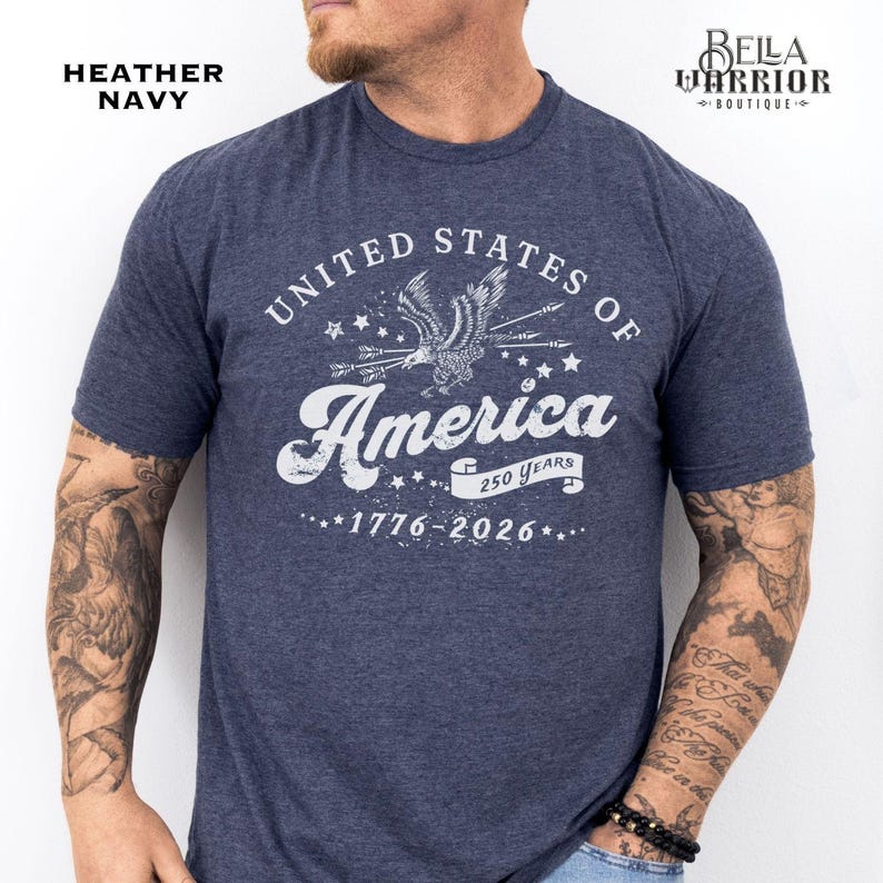 Puede incluir: Camiseta azul marino jaspeada con un dise&ntilde;o gr&aacute;fico blanco. El dise&ntilde;o incluye el texto "United States of America", un &aacute;guila, flechas y las fechas "1776-2026". Tambi&eacute;n incluye el texto "250 Years". Se ve el logo de Bella Warrior Boutique.