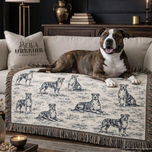 Op de afbeelding: Een gestroomde en witte hond ligt op een crèmekleurige sprei met een herhaald patroon van hondenillustraties. De sprei heeft een gefranjerde rand. Een kussen met de tekst "BELLA WARRIOR BOUTIQUE" is zichtbaar.