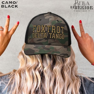 Könnte beinhalten: Camo- und schwarze Trucker-Mütze mit dem Text "FOXTROT DELTA TANGO" in goldfarbenen Buchstaben. Die Mütze hat einen Mesh-Rücken und einen gebogenen Schirm. Die Mütze wird von einer Person mit blonden Haaren getragen.