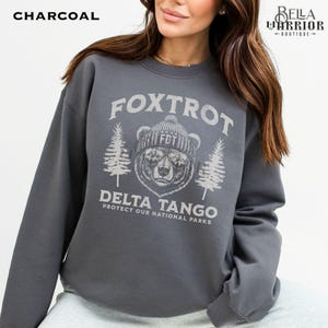 Puede incluir: Una sudadera gris carb&oacute;n con un gr&aacute;fico de un oso con un sombrero y gafas de sol. El texto "Fox Trot Delta Tango Protect Our National Parks" est&aacute; impreso en la sudadera.
