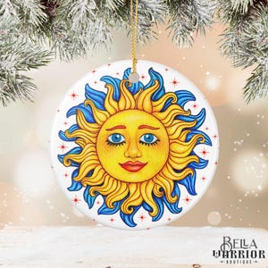 Może przedstawiać: Okrągła, biała ceramiczna ozdoba z uśmiechniętym słońcem. Słońce ma żółte i niebieskie detale, niebieskie oczy i czerwone usta. Czerwone gwiazdki są rozrzucone wokół słońca. Złoty sznurek jest przymocowany do zawieszenia.