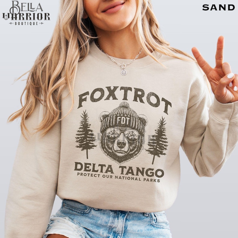 Puede incluir: Una sudadera beige con un gr&aacute;fico marr&oacute;n de un oso con gafas de sol y un sombrero con el texto "FDT" en &eacute;l. El texto "Fox Trot Delta Tango Protect Our National Parks" est&aacute; impreso en la sudadera.