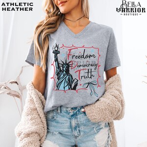 May include: Athletic heather gray V-neck t-shirt featuring a graphic of the Statue of Liberty holding a torch, with the words "Freedom, Democracy, Truth". The shirt has a red and blue design and is from Bella Warrior Boutique.