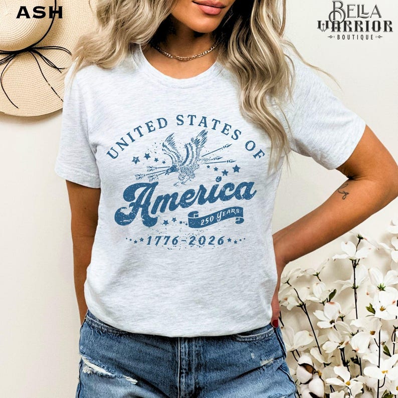 Puede incluir: Camiseta gris claro con un gr&aacute;fico azul que dice "United States of America" con un &aacute;guila y estrellas. El texto "250 Years 1776-2026" tambi&eacute;n est&aacute; impreso en la camiseta. La camiseta es de manga corta.
