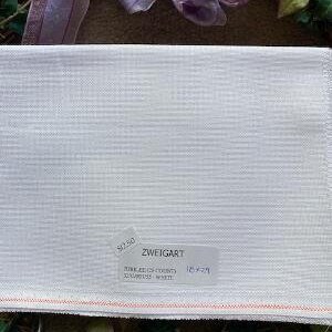 Könnte beinhalten: Weißer Zweigart-Stoff mit strukturiertem Gewebe. Der Stoff hat ein Etikett mit dem Markennamen und anderen Details. Die Kanten sind mit einer roten Naht versehen. Der Stoff ist von Grün und einem dekorativen Band umgeben.