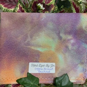 Puede incluir: Un trozo de tela teñido a mano en tonos de morado, rosa, naranja y verde. La tela está etiquetada con "Hand Dyes By Jim", "Lugana 32 count" y "Bewitched".