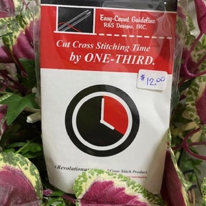 May include: A cross-stitch product package with a red and white label. The label reads "Cut Cross Stitching Time by ONE-THIRD" and includes a clock graphic. The text "Easy-Count Guideline" is also visible. The package is set against a backdrop of green and purple foliage.