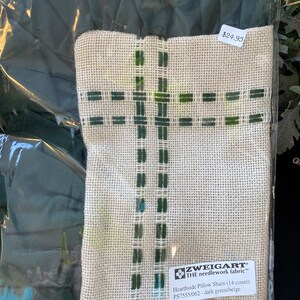 Puede incluir: Una funda de almohada beige con bordado verde oscuro en un patrón de rejilla. La funda está empaquetada en plástico y etiquetada con la marca Zweigart, "Hearthside Pillow Sham (14 count)", y "PS7555/062 - verde oscuro/beige". La etiqueta de precio dice "24,95 €".