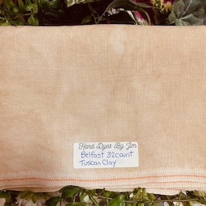 May include: A piece of hand-dyed linen fabric in a light brown color. The fabric is labeled "Hand Dyes By Jim Belfast 32 count Tuscan Clay."