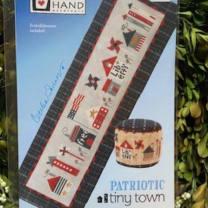 May include: A cross stitch pattern for a patriotic themed needlepoint project called "Patriotic Tiny Town". The pattern features a row of small houses with red, white, and blue accents. The pattern is printed on a blue background with a white border. The pattern is $13.00.