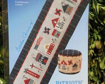 May include: A cross stitch pattern for a patriotic themed needlepoint project called "Patriotic Tiny Town". The pattern features a row of small houses with red, white, and blue accents. The pattern is printed on a blue background with a white border. The pattern costs £13.00.