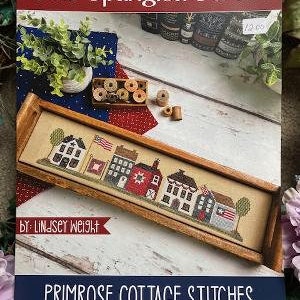 May include: A cross-stitch pattern featuring a row of houses with red, white, and blue accents. The pattern is titled "Star Spangled Street" and is by Lindsey Weight. The pattern is from Primrose Cottage Stitches.