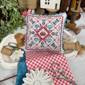 May include: A handmade cross-stitch pincushion with a star and sewing-themed design, surrounded by blue scalloped trim. The pincushion rests on a red and white checkered fabric, with a pair of silver scissors and wooden spools nearby. A craft-themed image.
