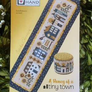 May include: A cross stitch pattern for a honeybee themed town with the text "A Honey of a tiny town". The pattern includes a small house with a beehive and sunflowers.