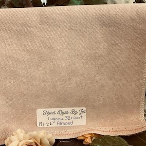 以下が含まれることがあります： 「Hand Dyes By Jim Lugana 32 count 18x26 Almond」と書いてあるラベルが付いた、薄いベージュの生地のアップ写真です。