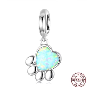 May include: A silver charm with a paw print design. The charm features a heart-shaped paw print with a light blue opal-like centre. The charm is made of 925 silver and has a loop for attaching to a bracelet or necklace.