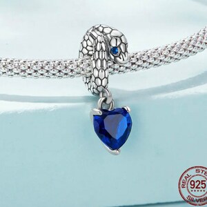 May include: A silver snake charm with a blue gemstone heart pendant. The snake has scale detailing and a blue eye. The heart-shaped gemstone is a deep blue colour. The charm is displayed on a silver chain.