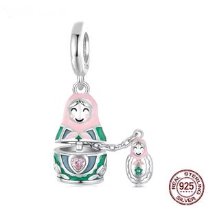 May include: A silver charm featuring a Russian nesting doll design. The main doll is pink and green, with a smaller doll attached by a chain. The charm has a heart-shaped gem and is marked "925 Silver".