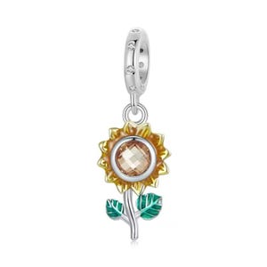 Pode incluir: Um berloque de prata com um design de girassol. O girassol tem uma flor amarela e laranja com uma pedra central facetada. O caule e as folhas são verdes. O berloque tem um laço com pequenas pedras transparentes.