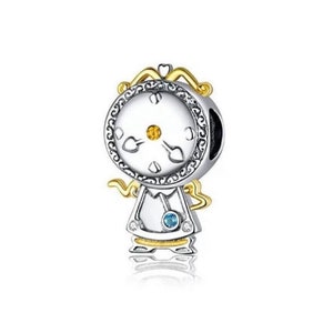Puede incluir: Un charm de plata con forma de reloj con detalles dorados. La esfera del reloj es blanca con manecillas negras y un centro dorado. El charm tiene una pequeña gema azul y está diseñado para parecerse a una persona.