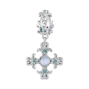 Pode incluir: Um berloque prateado com um pendente de cruz. A cruz apresenta uma pedra central azul clara e é adornada com pedras preciosas menores, azul claro. O berloque tem um design ornamentado com detalhes florais e um anel decorativo.