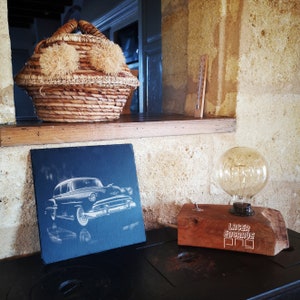 May include: A black slate plaque with a white etching of a vintage car. The plaque is leaning against a wooden surface. A wooden lamp base with a light bulb and the text "Laser Engrave Pro" is on the surface.