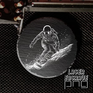 Puede incluir: Un posavasos de pizarra negra con un dise&ntilde;o grabado con l&aacute;ser blanco de un snowboarder. El dise&ntilde;o incluye un snowboarder en el aire, una tabla de snowboard y un paisaje de monta&ntilde;a nevado. El posavasos es redondo y tiene una superficie lisa. El texto "LASER ENGRAVE" es visible en la esquina inferior derecha de la imagen.