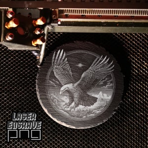 Puede incluir: Un posavasos de pizarra negra con una imagen grabada con l&aacute;ser de un &aacute;guila calva en vuelo. El &aacute;guila vuela sobre una cordillera con nubes en el fondo. La imagen est&aacute; rodeada por un c&iacute;rculo.